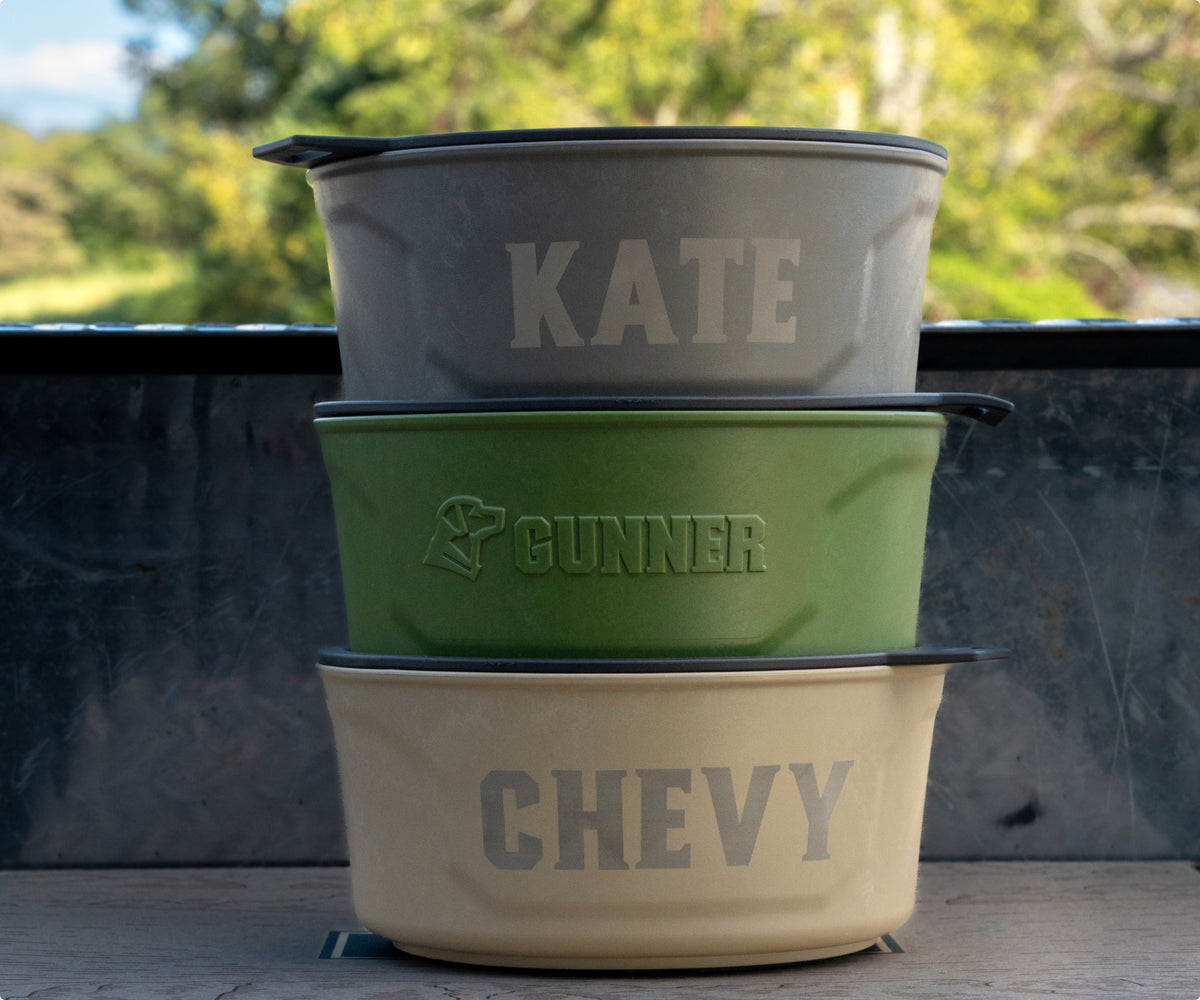 Three metal pet bowls are stacked outdoors, labeled “KATE” (gray), “GUNNER” (green) with a logo, and “CHEVY” (cream)—perfect for use at your dog kennel or beside the hunting dog crate, with trees in the background.