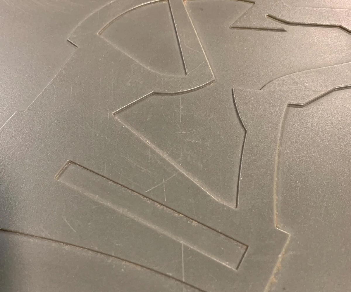 Close-up of a metallic surface with an engraved outline of a stylized sword, helmet, and a hunting dog, possibly representing a knight or warrior in simple, angular lines.