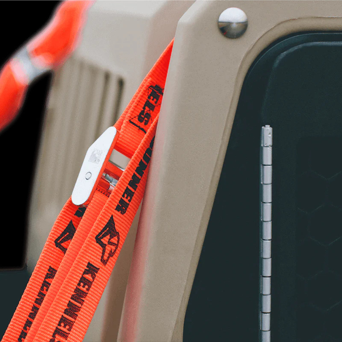 Close-up of an orange Key Skner-branded strap attached to a beige dog crate, with a metal clasp securing the strap and part of the carrier’s black door visible on the right.