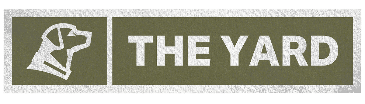 A rectangular green sign with a white outline, featuring a simple working dog head silhouette on the left and the bold white text THE YARD on the right.