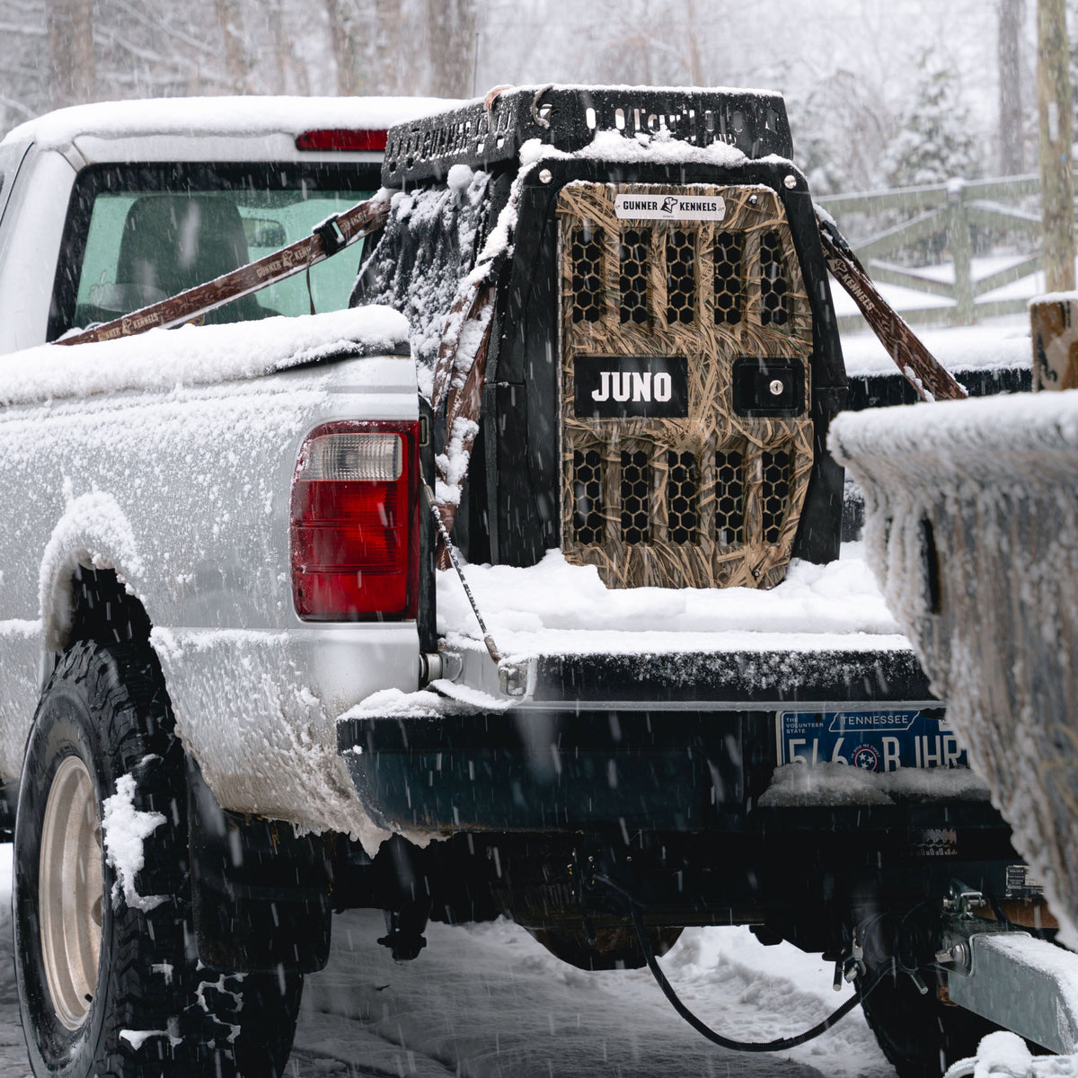 A snow-covered pickup with a GUNNER G1™ KENNEL - BLACK/SHADOW GRASS, labeled Juno, secured in the bed. Snow falls as a Tennessee plate peeks through, with trees and a railing visible in the background.