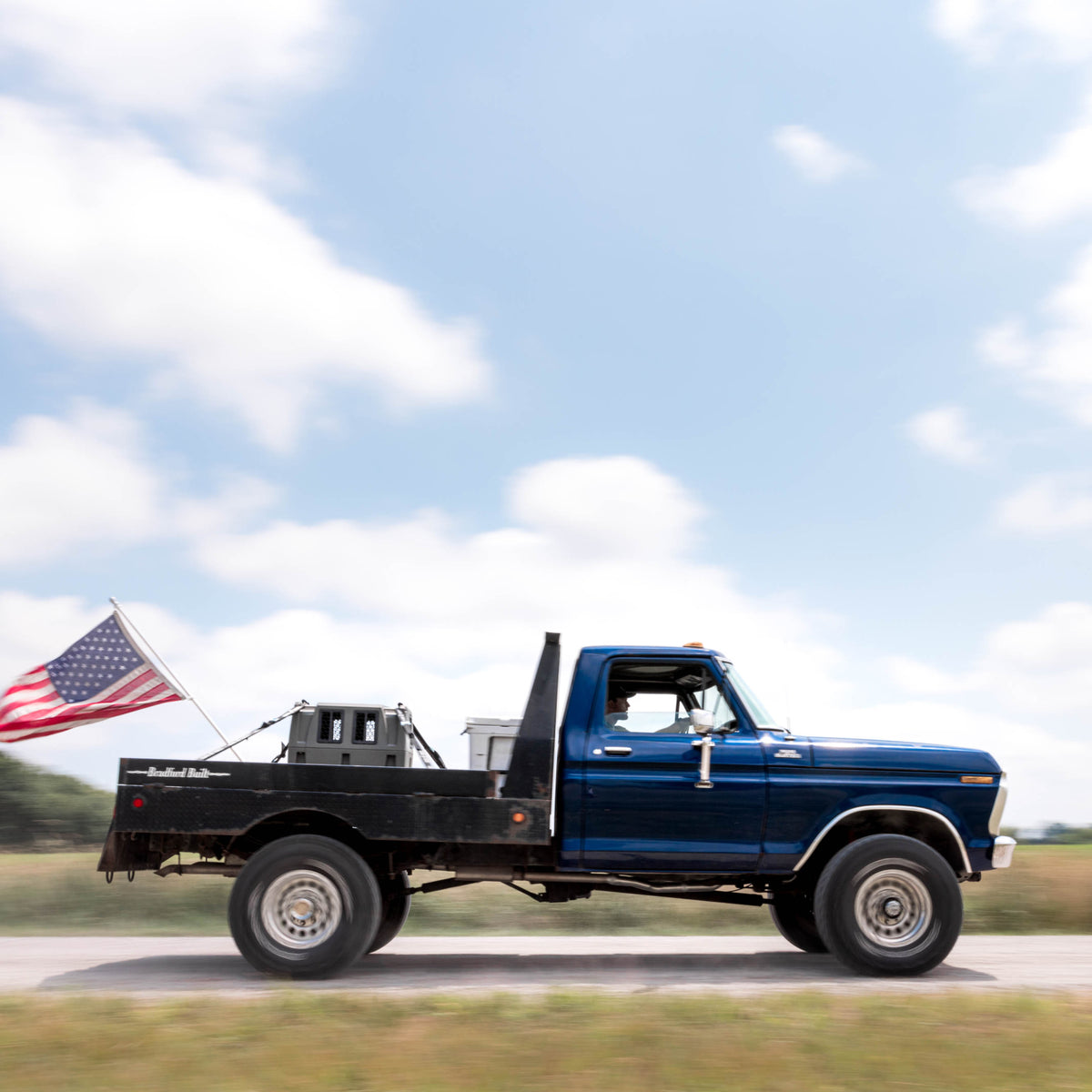 A blue pickup with a flatbed and an American flag drives down a rural road under partly cloudy skies, carrying a GUNNER G1™ KENNEL - GUNMETAL securely in the bed for loyal companions.