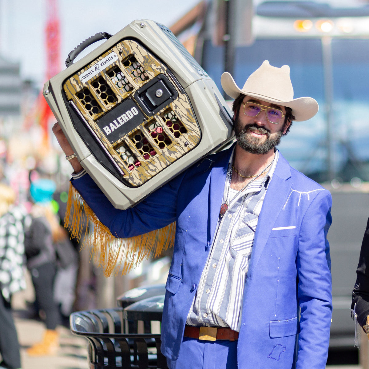 A man in a light tan cowboy hat, glasses, and a blue suit with a bolo tie smiles while carrying a GUNNER G1™ KENNEL - MOSSY OAK BOTTOMLAND GUNMETAL on his shoulder like its the toughest crate. A bus and people are visible in the blurred background.