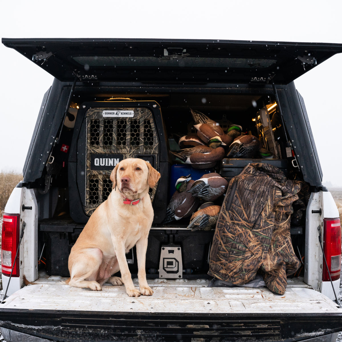 A yellow labrador sits on the open tailgate of a white truck, surrounded by hunting gear and decoys, with the GUNNER G1™ KENNEL - BLACK/SHADOW GRASS dog crate ready in the snowy truck bed—perfect for duck & goose hunters.