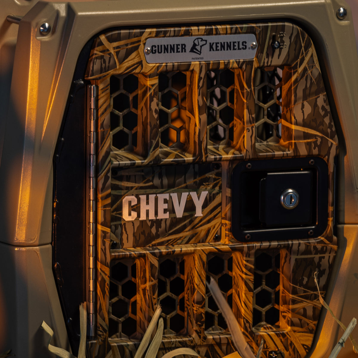 Close-up of a GUNNER G1™ KENNEL - TAN/SHADOW GRASS crate door labeled CHEVY, with a metal GUNNER plate above. Dry grass can be seen at the bottom of the image.