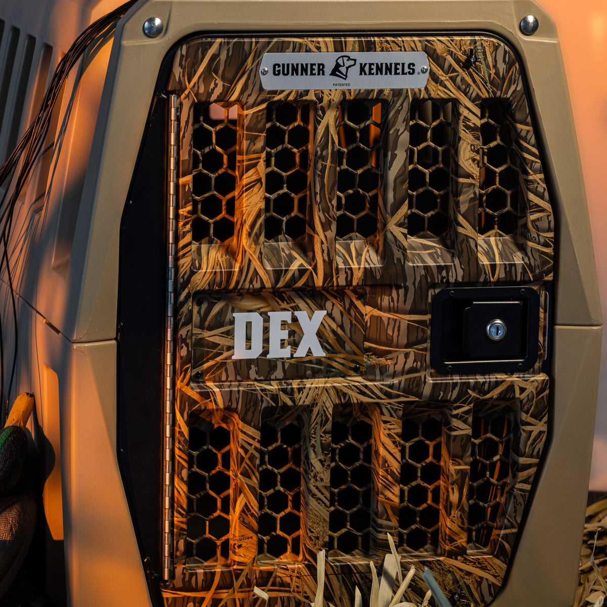 A close-up of the GUNNER G1™ KENNEL - TAN/SHADOW GRASS with a metal lock, set among dry grass and outdoor gear—ideal for adventurers seeking the safest dog crate for rugged trips.
