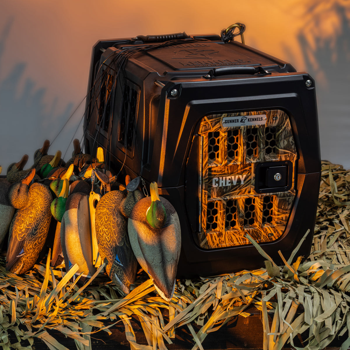 A GUNNER G1™ KENNEL in Black/Shadow Grass stands on straw beside vibrant duck decoys, illuminated by warm light.