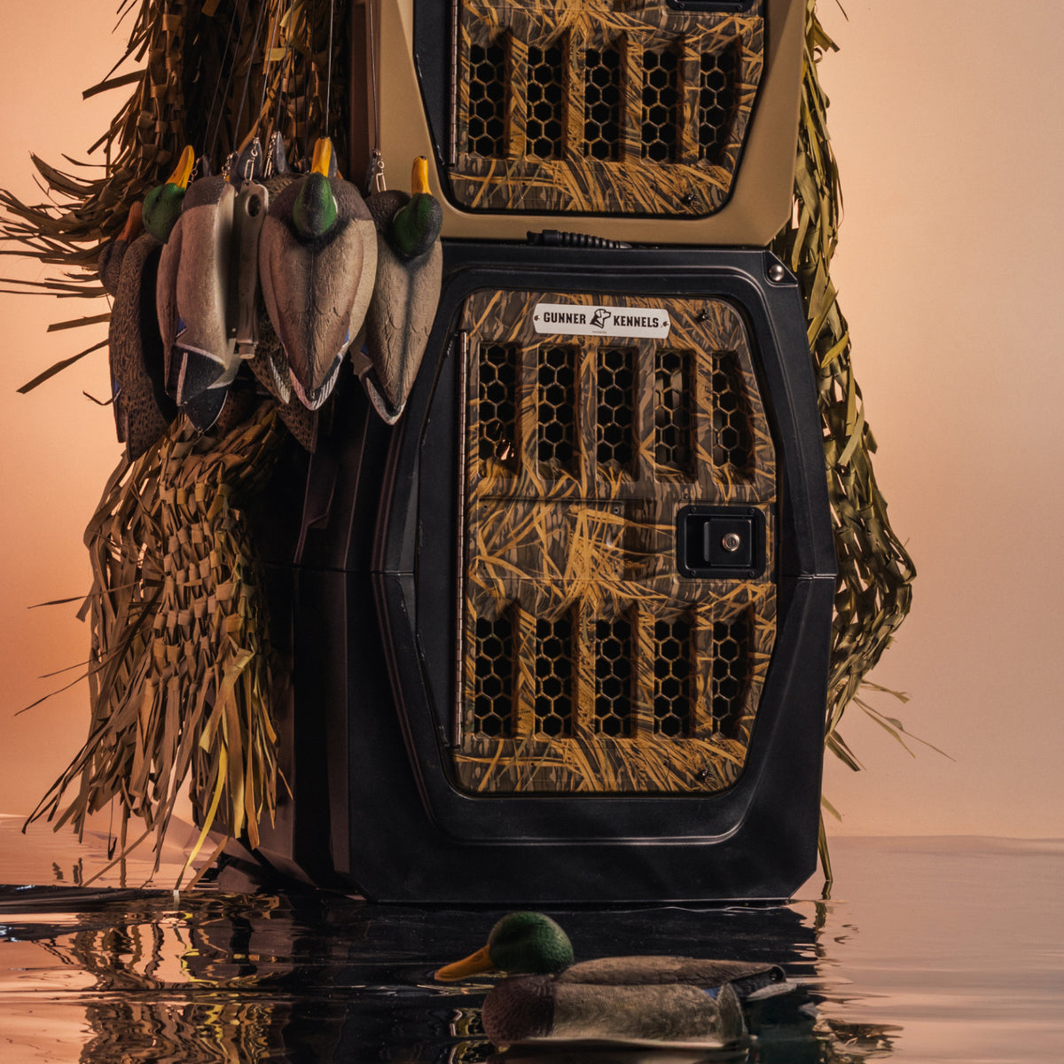 A stack of GUNNER G1™ KENNEL - BLACK/SHADOW GRASS kennels, adorned with duck decoys and grass accents, reflected in water against a beige backdrop—ideal for duck hunters.