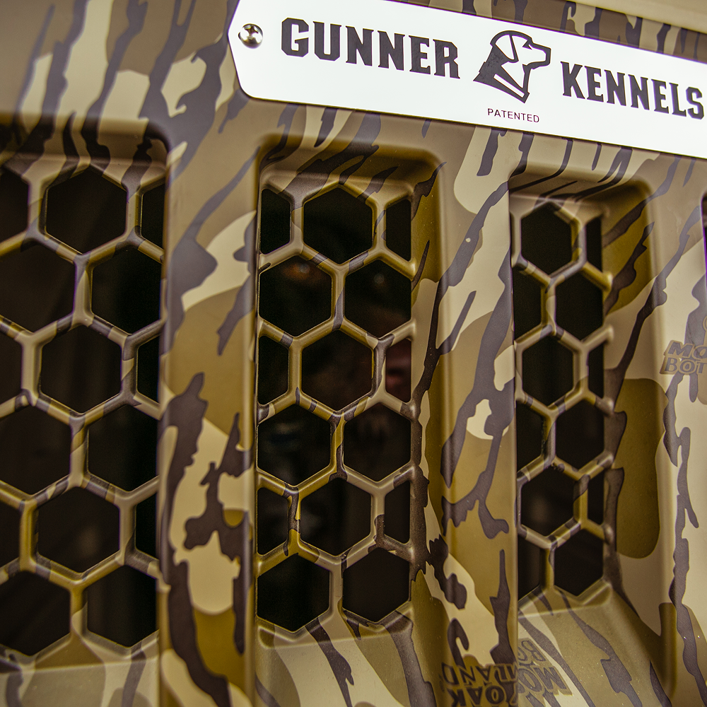 Close-up of the GUNNER G1™ KENNEL - MOSSY OAK BOTTOMLAND GUNMETAL, showcasing its camouflage pattern, honeycomb ventilation grilles, and a branded metal nameplate at the top.