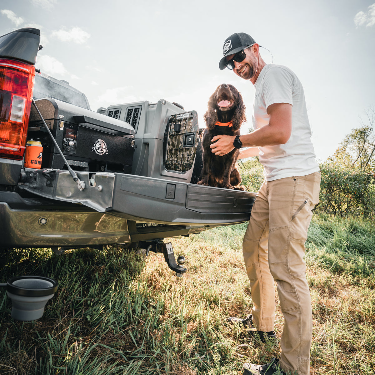 A man in sunglasses and a cap smiles by an open truck tailgate, petting a brown dog beside the GUNNER G1™ KENNEL - MOSSY OAK BOTTOMLAND GUNMETAL. Its a sunny day outdoors with grass and trees in the background.