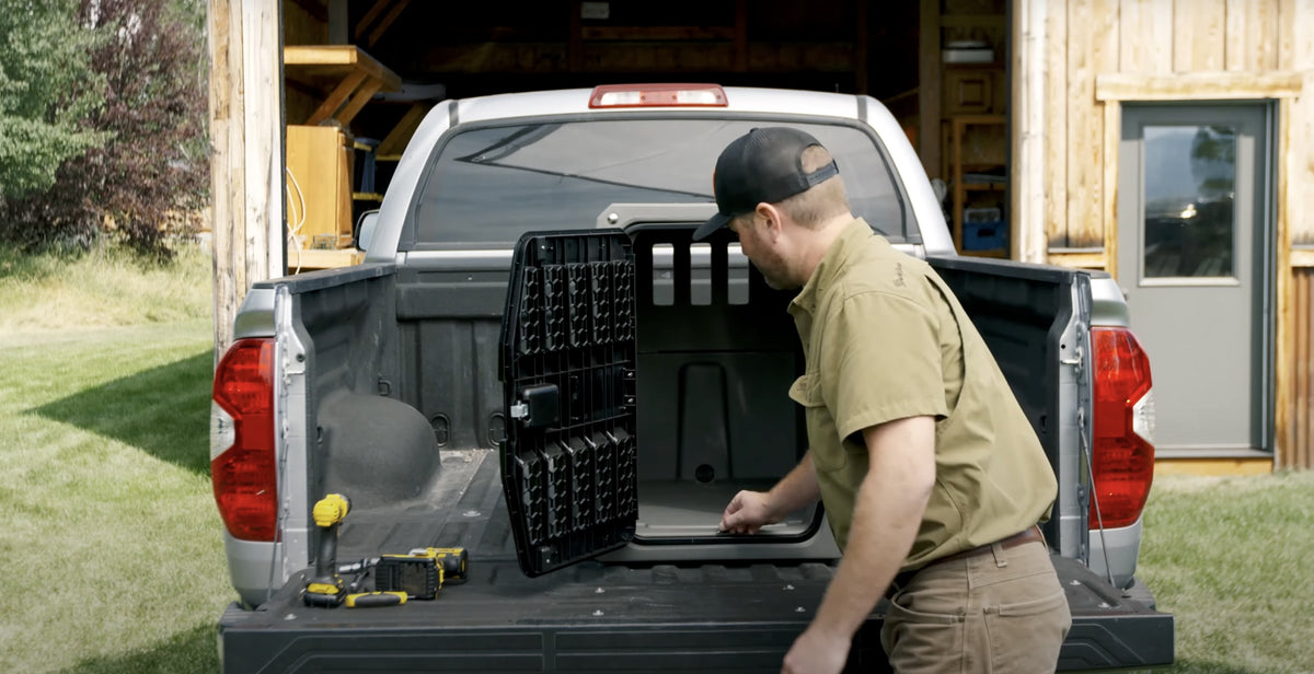 A man wearing a cap and khaki shirt installs a black storage unit—perfect for hunting dog or working dog gear—in the bed of a white pickup truck, with tools and a drill nearby. The truck is parked outside a wooden garage or workshop.