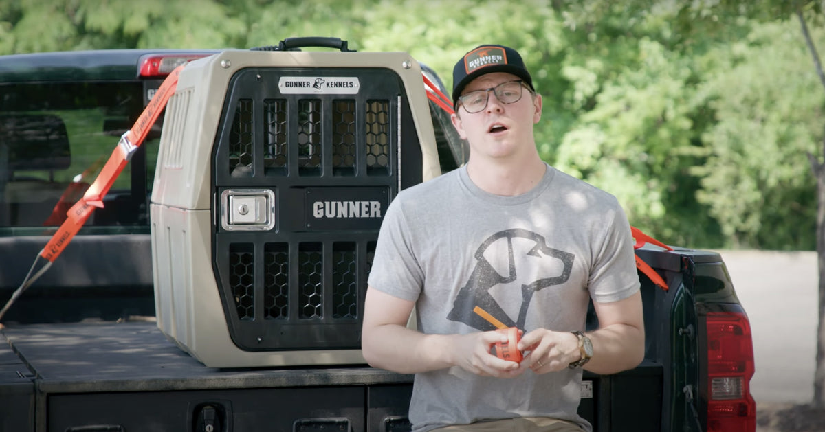 A person wearing glasses, a cap, and a dog-printed t-shirt sits on the back of a truck beside a large GUNNER dog crate, perfect for any hunting dog, with a green, tree-filled background.