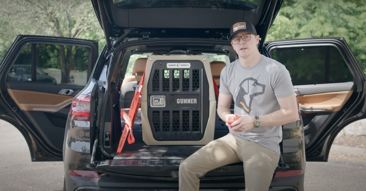 A person wearing glasses, a hat, and a t-shirt sits on the edge of an SUV’s open trunk next to a large, secured dog crate labeled Gunner. Trees and greenery are visible in the background.