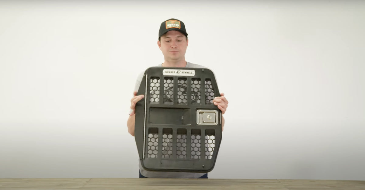 A person wearing a black cap and gray shirt holds up a black, vented dog kennel door with a metal latch, standing behind a wooden table against a plain white background.