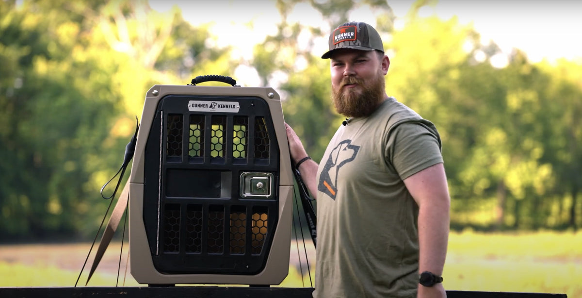 A bearded man in a cap and t-shirt stands outdoors, smiling while resting his hand on a large, heavy-duty dog kennel. Trees and sunlight create a blurred green background—perfect for any hunting dog or working dog.