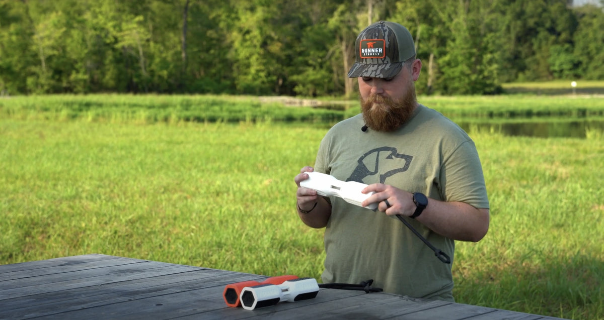 A bearded man wearing a cap and an olive green shirt stands outdoors at a wooden table, examining a white electronic device—part of his outdoor gear—with another similar device and green trees visible in the background.