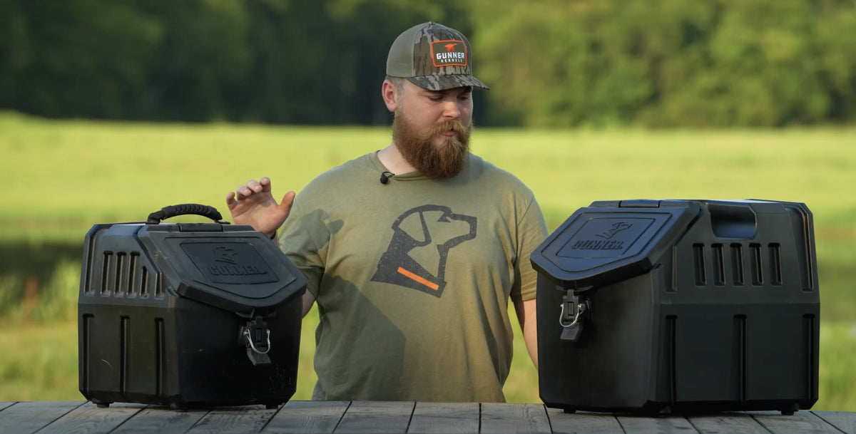 A bearded man in an olive T-shirt with a dog graphic and a baseball cap stands outdoors behind a table, comparing two black Bunker dog kennel storage containers. Green trees and grass are in the background.