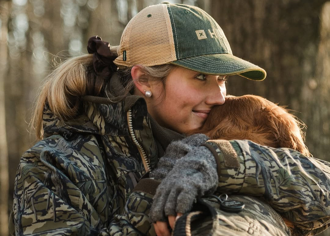 A woman in camouflage clothing and a green cap hugs her brown hunting dog outdoors, both looking content. Trees are visible in the blurred background.