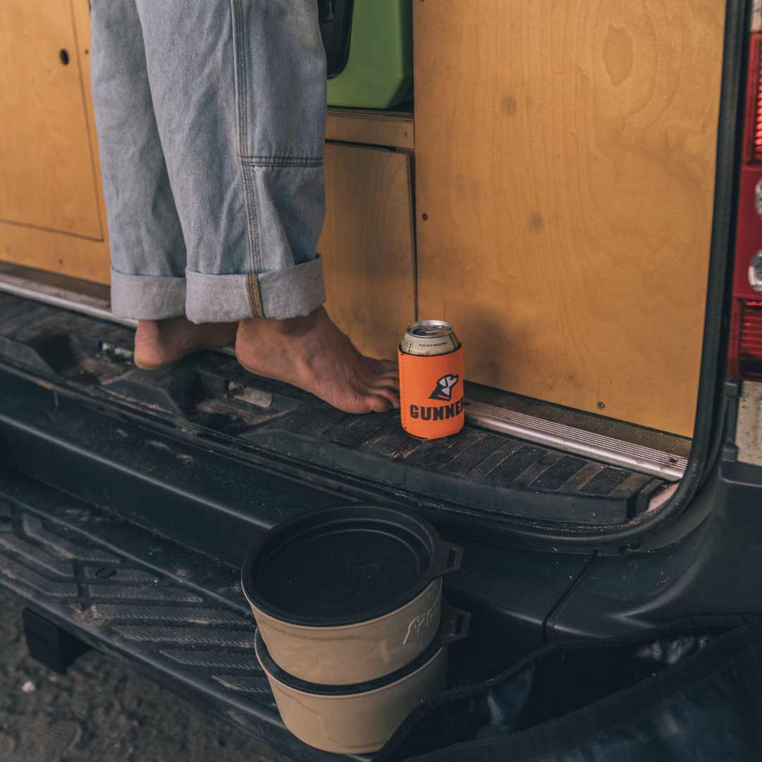 A person with rolled-up jeans stands barefoot on a vans back. Nearby are an orange drink can in a GUNNER Koozie Pack and two stacked containers used for kennel supplies for their working dog.