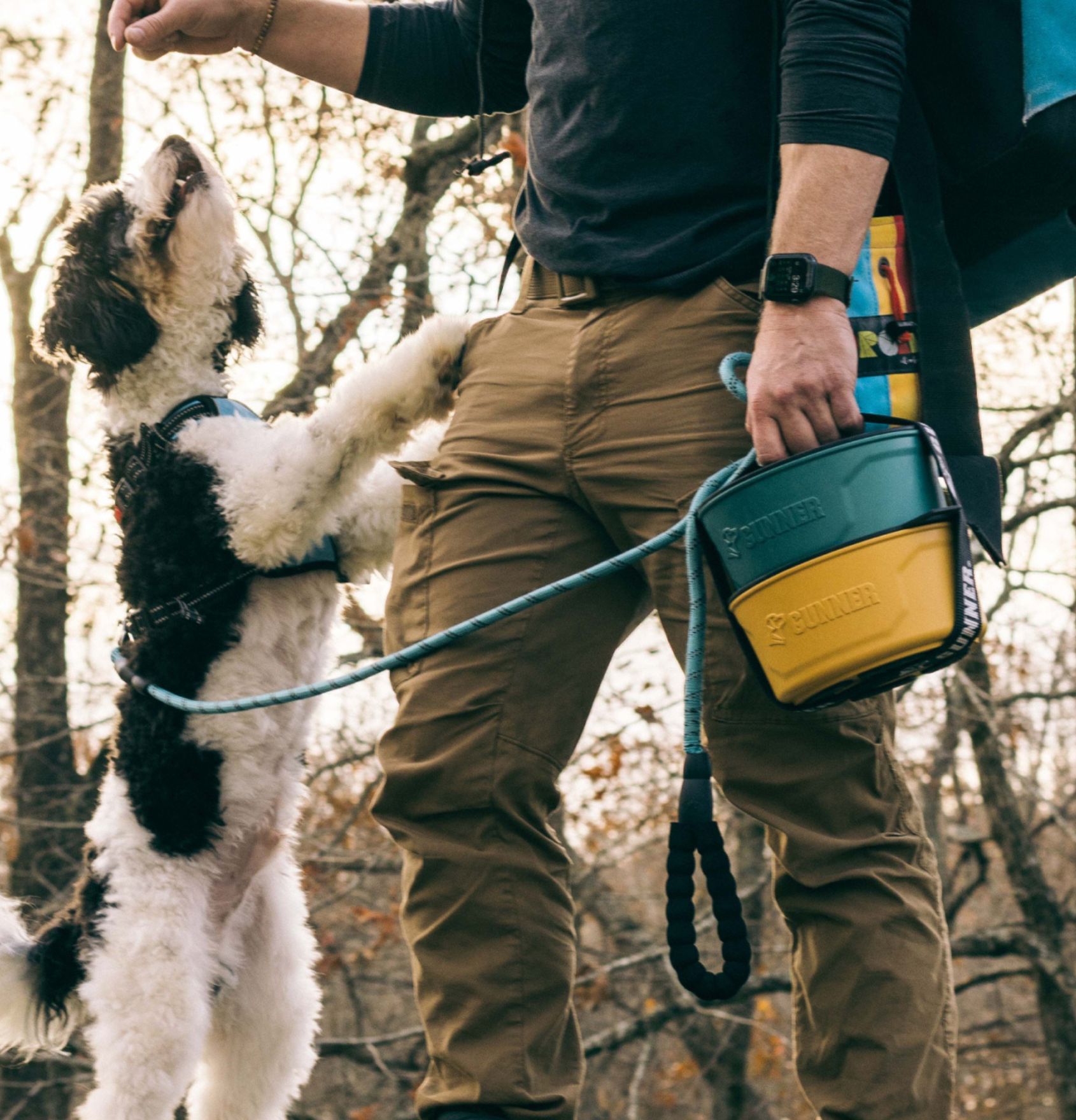 A person standing outdoors, dressed in khaki pants and outdoor gear with a smartwatch, holds a leash and colorful container while giving a treat to a black and white working dog standing on its hind legs. Trees are visible in the background.