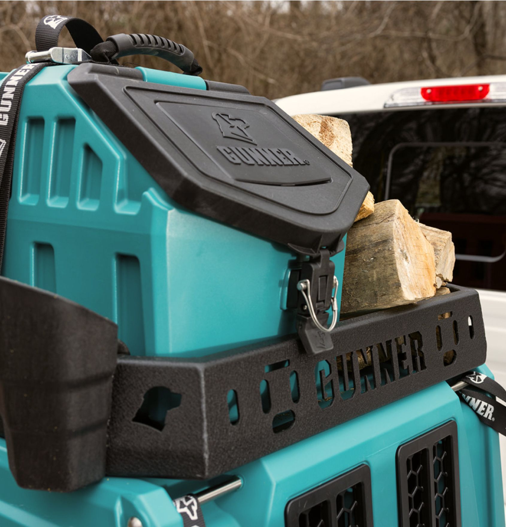 Close-up of a teal GUNNER storage box, perfect for organizing outdoor gear, secured in the back of a vehicle with split firewood stacked on top. The scene is outdoors near some trees.