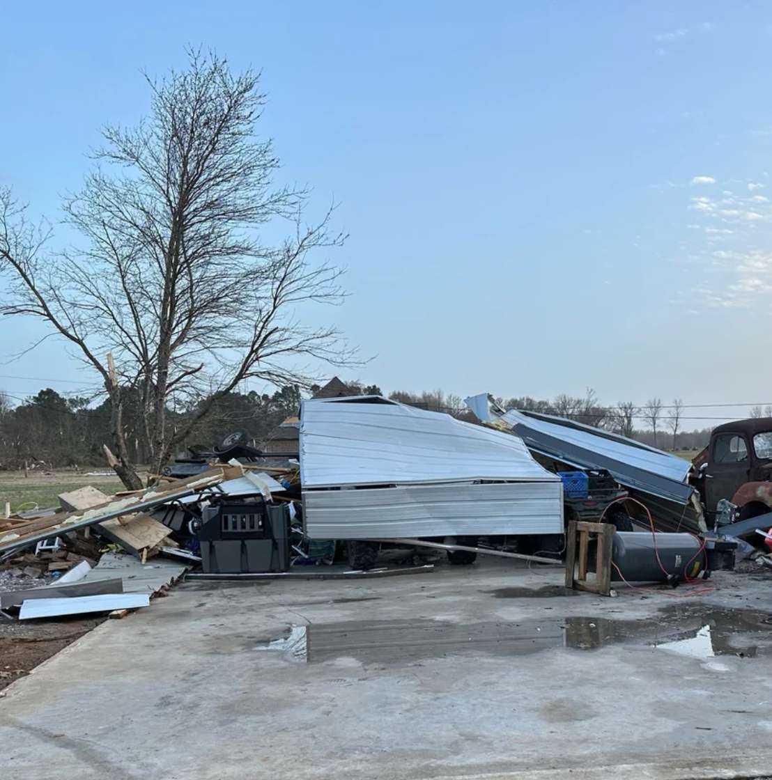 Debris from a collapsed building, including metal panels and wood, is scattered across a concrete slab. A bare tree stands nearby and a vehicle with a dog crate in the back is partially visible on the right under a blue sky.