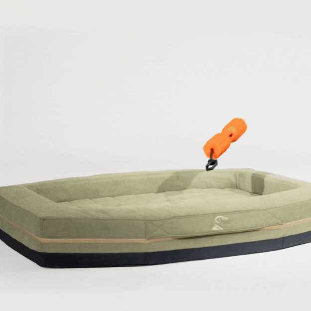 A bright orange dog toy attaches to a khaki-colored dog bed with raised edges and a black base, showing how the toy clips onto the bed—an ideal setup for your hunting dog’s crate. The background is plain white.