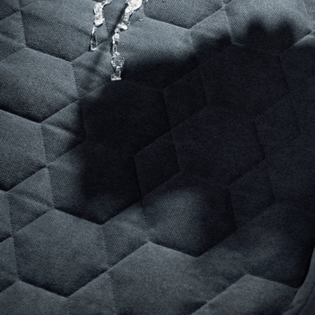 A close-up of water droplets on a dark, hexagon-patterned quilted fabric surface, reminiscent of premium kennel bedding designed for a hunting dog, with shadows and light highlighting the texture.