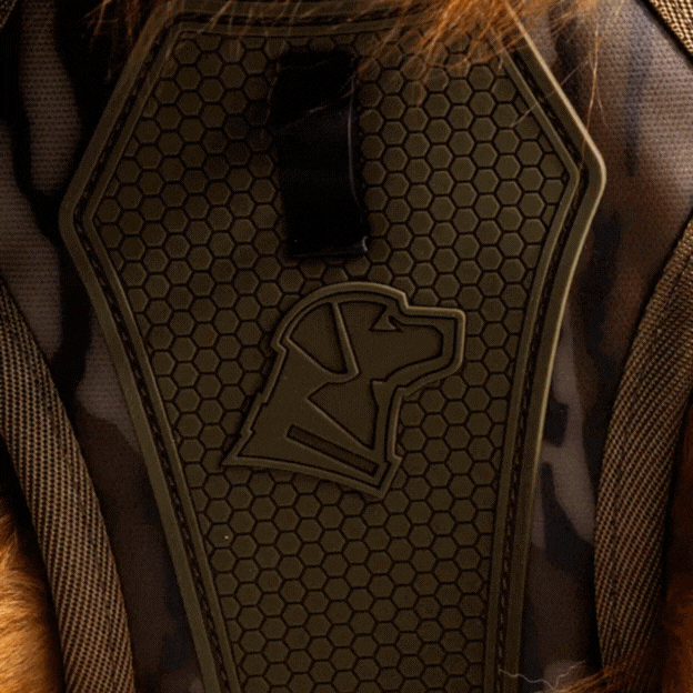 Close-up of a camouflage-patterned dog harness with a hexagon texture, perfect for any hunting dog, and an outline of a dogs head embossed on the chest plate. Brown fur is visible around the edges.