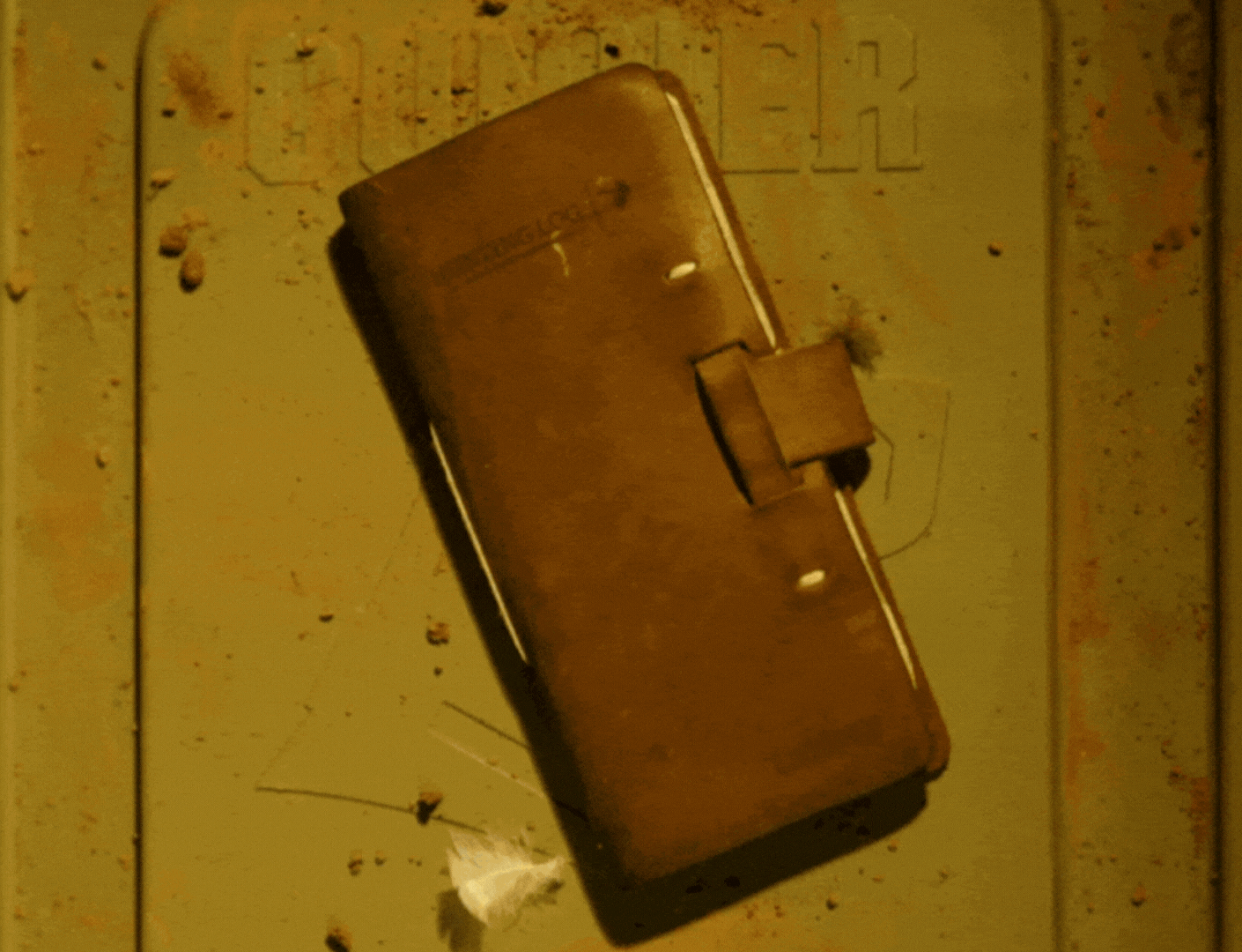 A brown leather wallet lies on a dusty, tan metal surface—like the top of a rugged dog crate—with small debris scattered around and the word GUNNER embossed in the background.