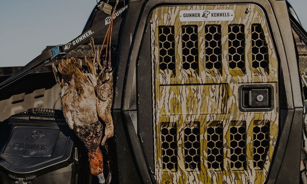 A camouflage-patterned Gunner Kennels crate is mounted on a vehicle, with three hunted ducks hanging on a strap beside it—perfect for any hunting dog. The scene is outdoors under clear skies.