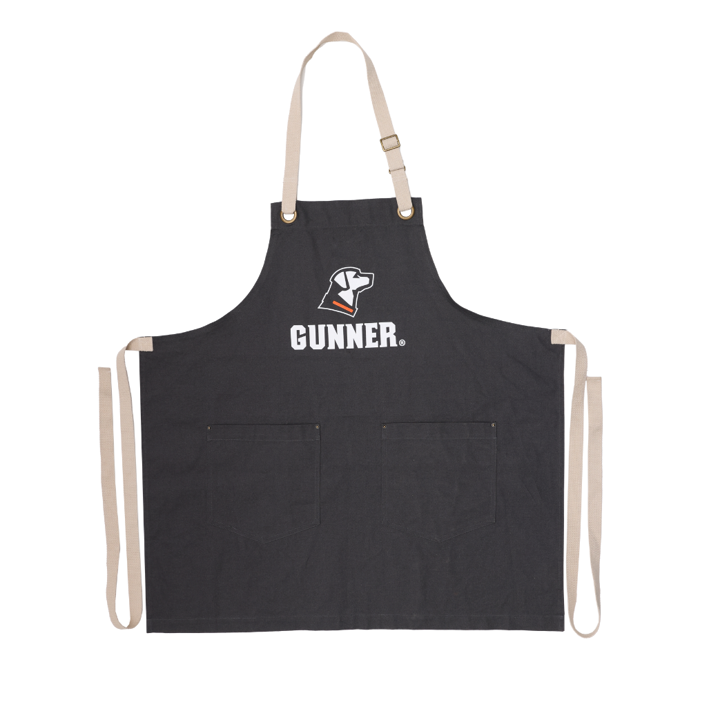 The black GUNNER Apron by GUNNER has beige neck/waist straps, a white dog head logo, “GUNNER” on the chest, and two front pockets—ideal for tailgates or crawfish boils.