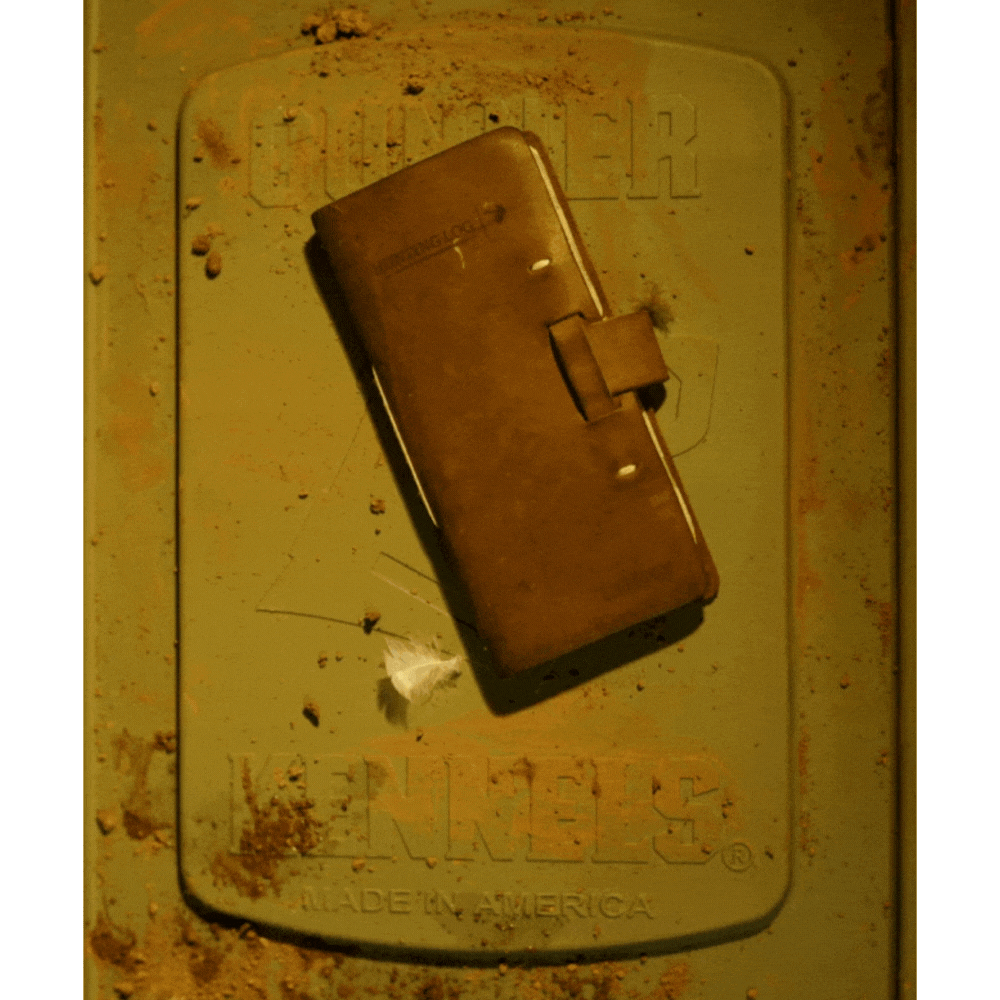 A GUNNER Hunting Log by GUNNER, styled like a limited-edition hunting journal, rests on a weathered green metal surface embossed with QUAKER KENNELS MADE IN AMERICA. A small plastic piece sits nearby.