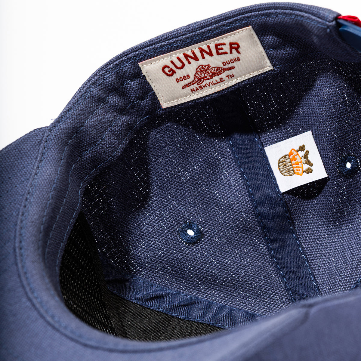 Close-up of the Kennel Patch Hat by GUNNER in navy blue, featuring a white label with red GUNNER DOGS DUCKS NASHVILLE, TN text and a small tag showing a cartoon dog next to a tree stump. Designed for anywhere you go.