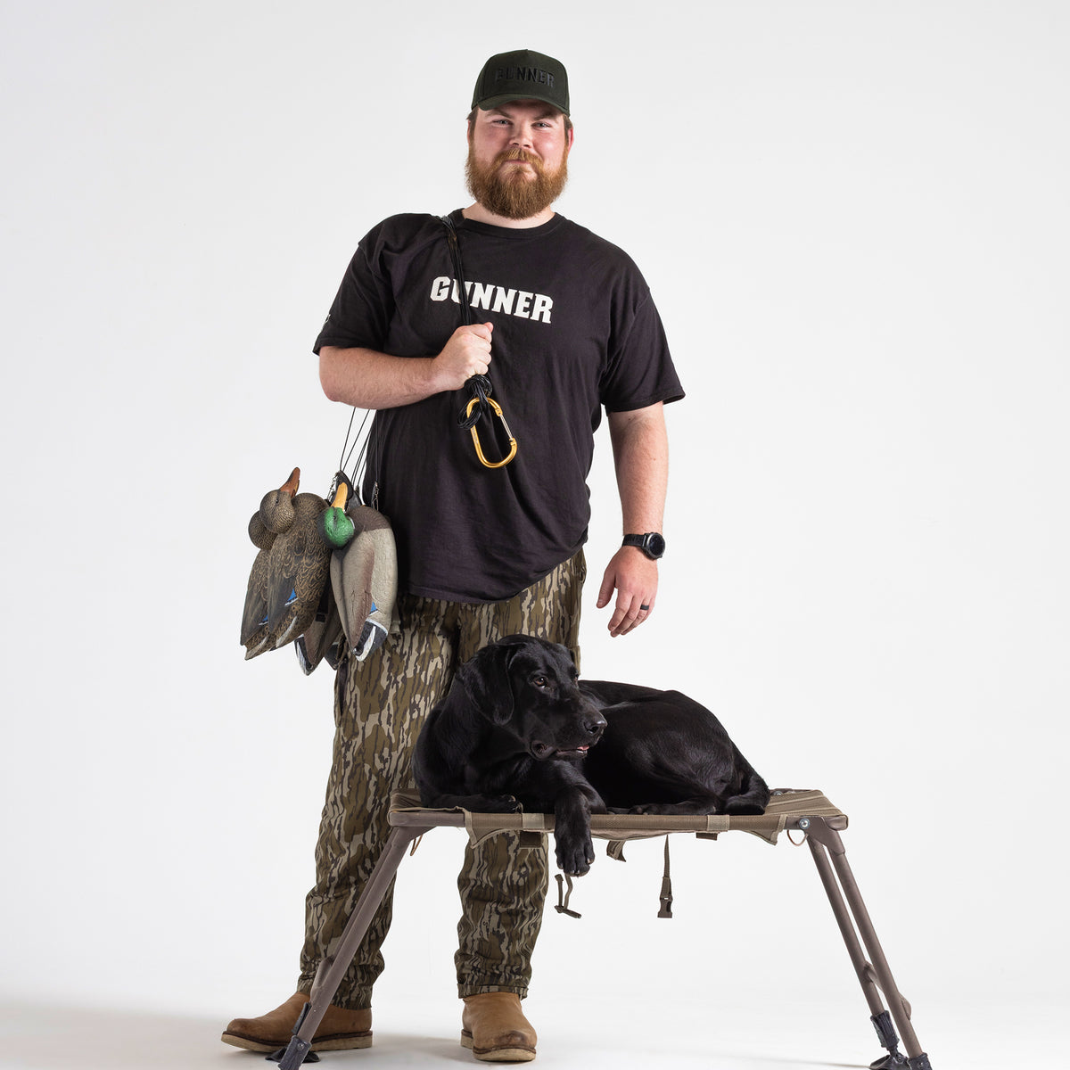A bearded man in camouflage pants, a GUNNER t-shirt, and the GUNNER Waxed Hat stands holding duck decoys. A black dog lies on a raised platform in front of him; both face the camera against a plain white background.