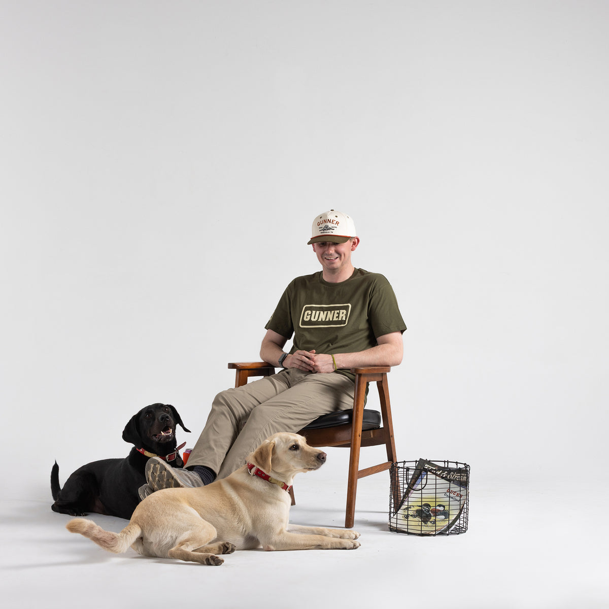 A man in a green GUNNER t-shirt and a white D & D Rope Hat by GUNNER with a green bill sits smiling on a wooden chair. Two dogs, one black and one yellow, sit beside him. Nearby is a black crate with a yellow cushion and rust-colored rope.
