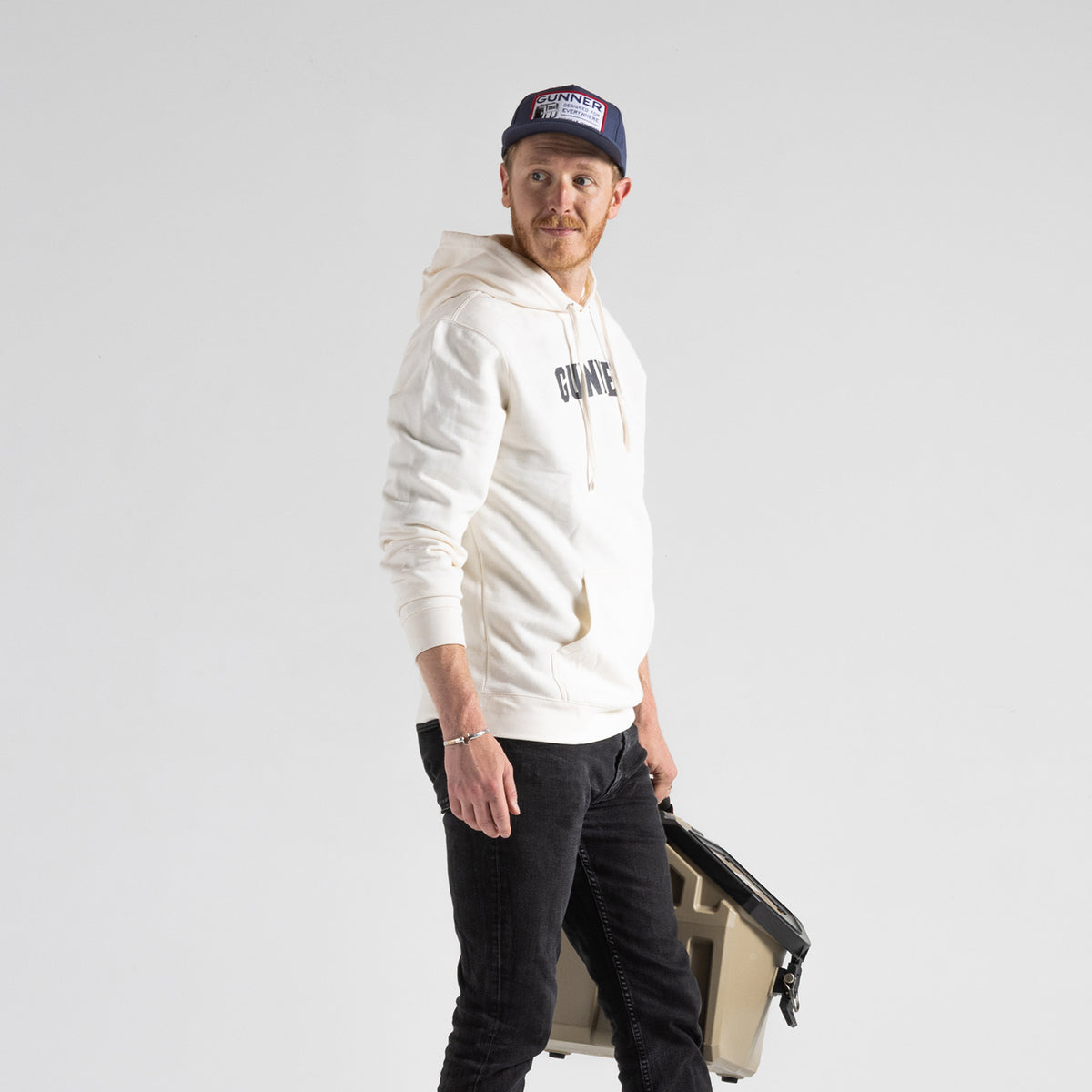 A man in a white hoodie and black jeans holds a tan cooler, wearing the GUNNER Kennel Patch Hat. He gazes slightly left against a plain white background, showcasing laid-back style suitable for any setting.