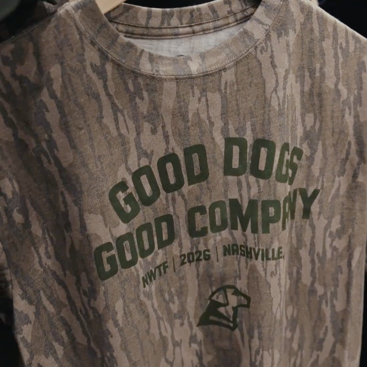 A Good Dogs, Good Tees shirt by GUNNER with a Mossy Oak Bottomland camo pattern hangs on a rack, displaying green text: GOOD DOGS GOOD COMPANY NWTF 2026 NASHVILLE and a small dog head logo below.
