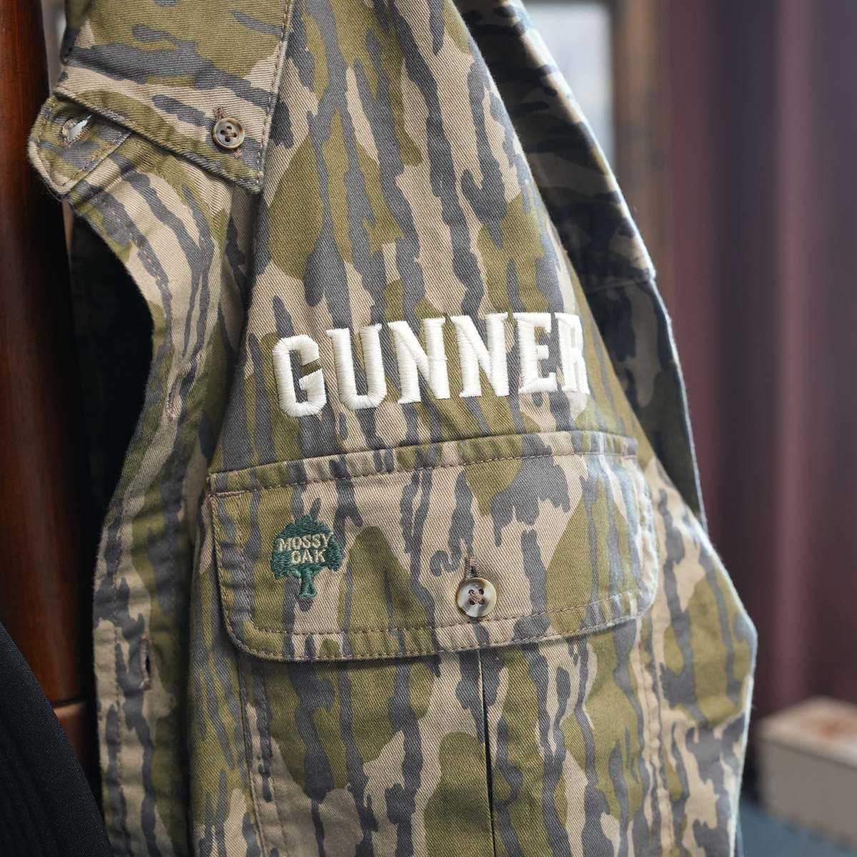 A camouflage GUNNER vest with a Mossy Oak Bottomland patch hangs on wood, ideal for pairing with the Late Season Long Sleeve Shirt Mossy Oak OG Bottomland by GUNNER.