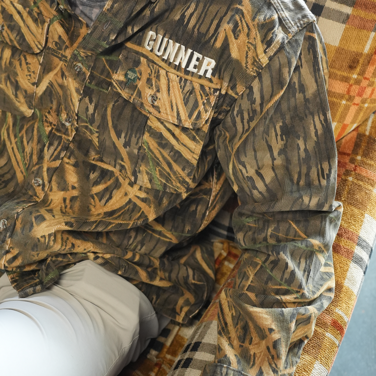 Someone wearing a GUNNER Late Season Long Sleeve Corduroy Mossy Oak OG Shadow Grass shirt is seated in a plaid chair, with only part of their torso, arm, and legs visible.