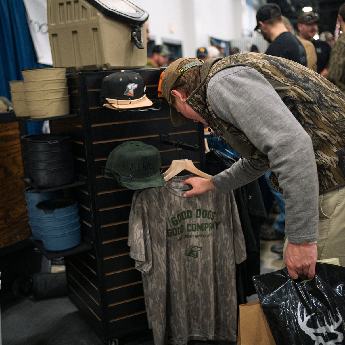 A man in a camouflage vest checks out a gray Good Dogs, Good Tees shirt by GUNNER at a retail display with hats and outdoor gear, as NWTF 2026 signage appears nearby.