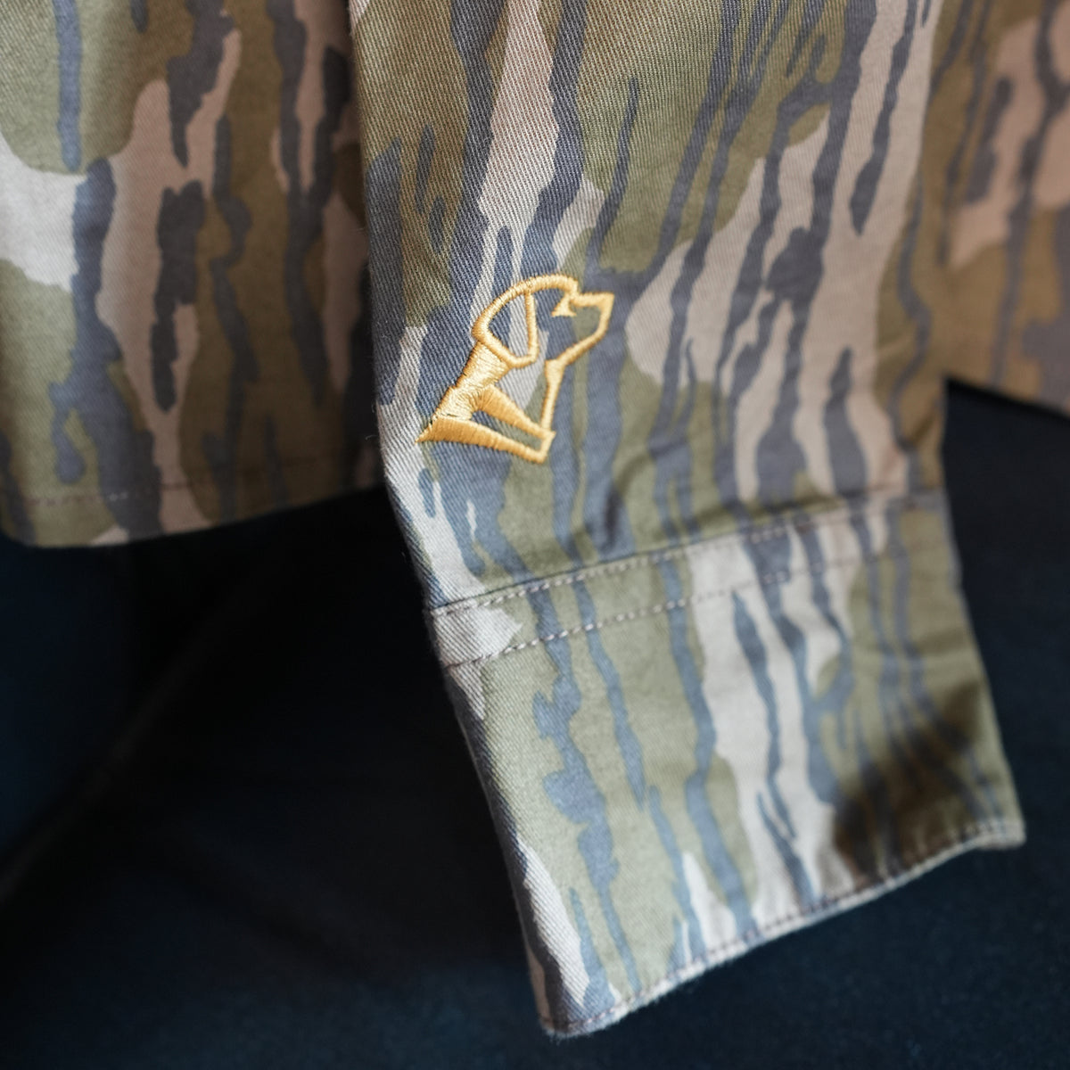 A close-up of the GUNNER Late Season Long Sleeve Shirt in Mossy Oak OG Bottomland camo shows its cotton twill sleeve and a small yellow embroidered detail near the cuff.