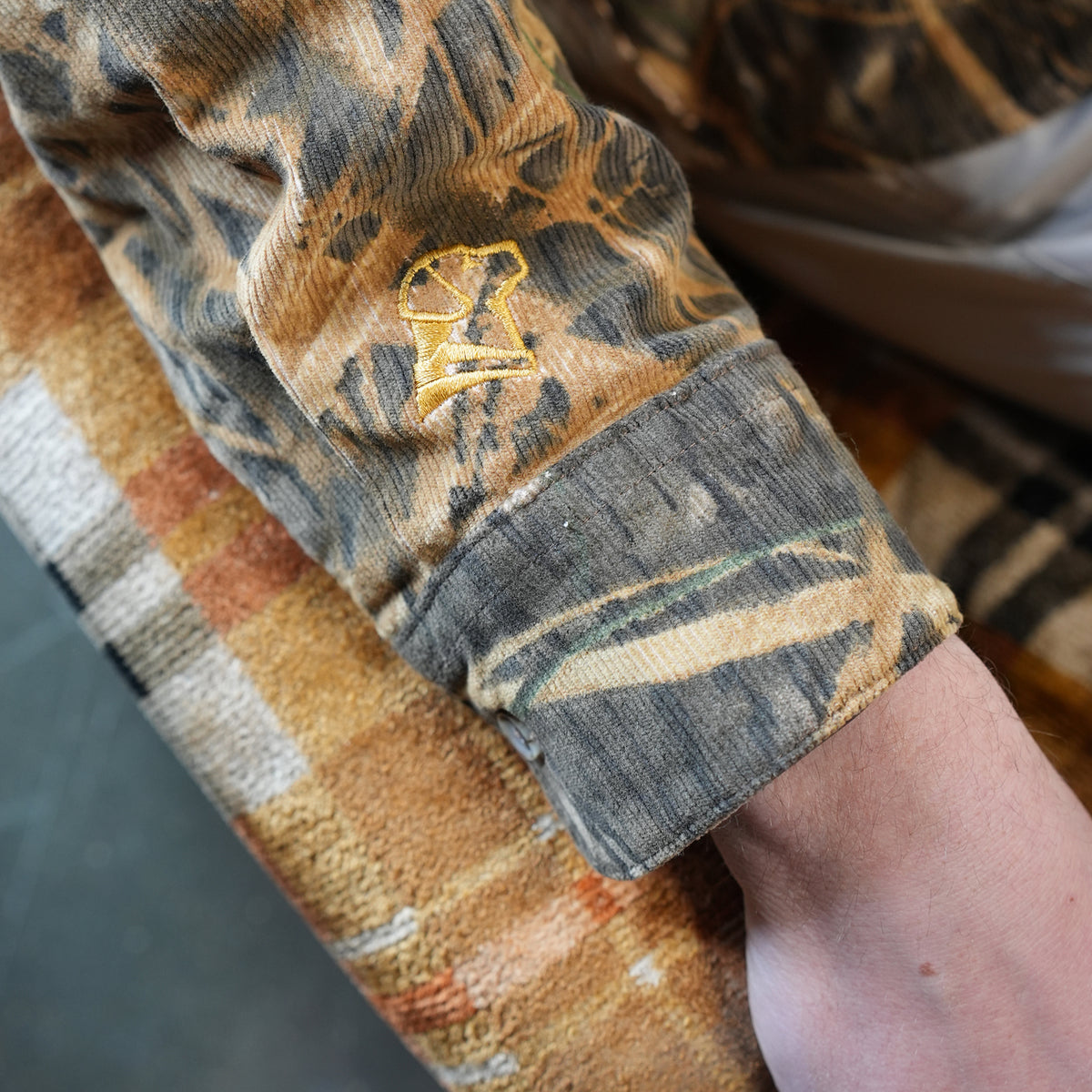 A close-up of a persons arm in the GUNNER Late Season Long Sleeve Corduroy Mossy Oak OG Shadow Grass jacket, featuring a yellow embroidered bird logo on the sleeve, resting on a brown and orange checkered corduroy shirt.