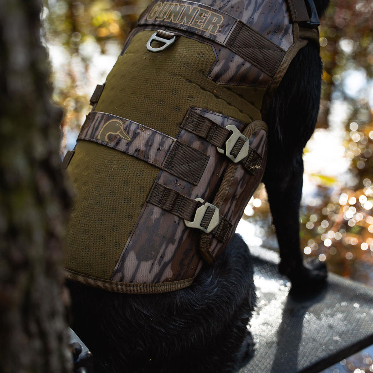 A black dog wearing a GUNNER Waterfowl Dog Vest Mossy Oak Bottomland® with a 5-Layer Performance System sits on an outdoor platform by a tree, with water and autumn foliage in the background.