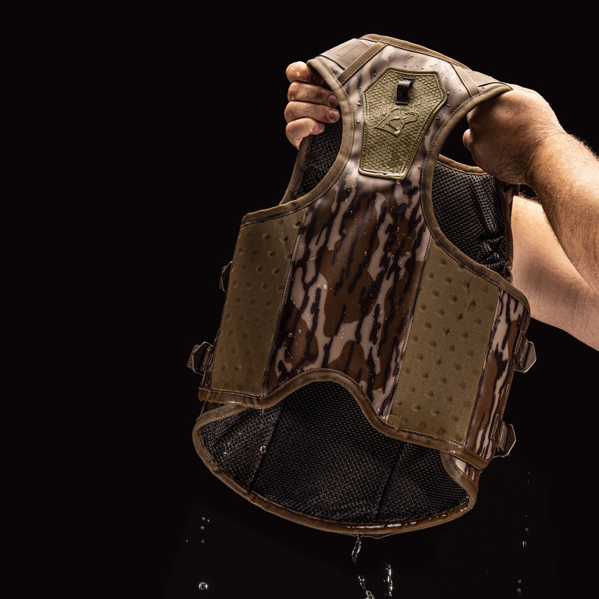 A person holds up a GUNNER Waterfowl Dog Vest Mossy Oak Bottomland® with both hands as water drips from its 5-Layer Performance System, set against a black background.