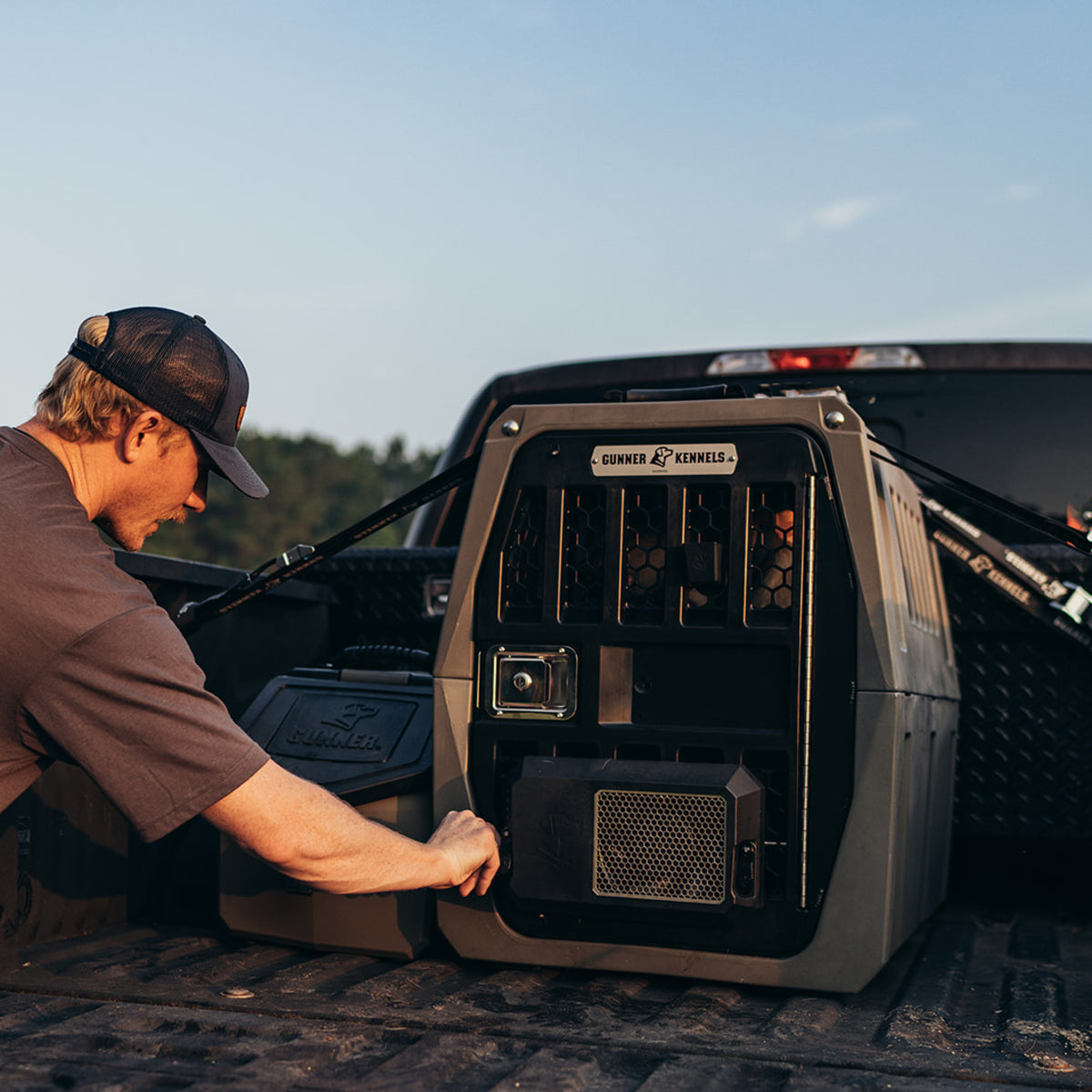 A person in a cap secures a large, rugged dog crate labeled Gunner Kennels in the back of a pickup truck bed at sunset. The kennel is fastened with straps for safe transportation, highlighting essential outdoor gear for pet owners.