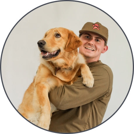 A smiling person in a brown cap and shirt holds a happy golden retriever, known for being an excellent hunting dog, in their arms against a plain light background.