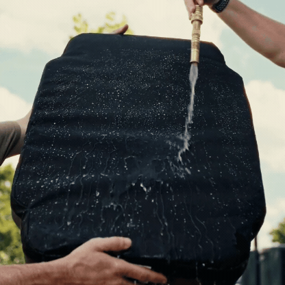 Two people hold a black cushion upright while someone sprays water on it with a hose, demonstrating that the water beads off the surface, indicating it is water-resistant—perfect for outdoor gear or use in a kennel. Trees and a blue sky are visible in the background.