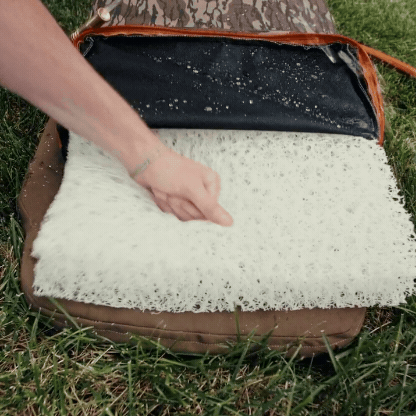 A hand lifts a white, porous foam pad from inside a zippered brown dog kennel case, resting on green grass outdoors.