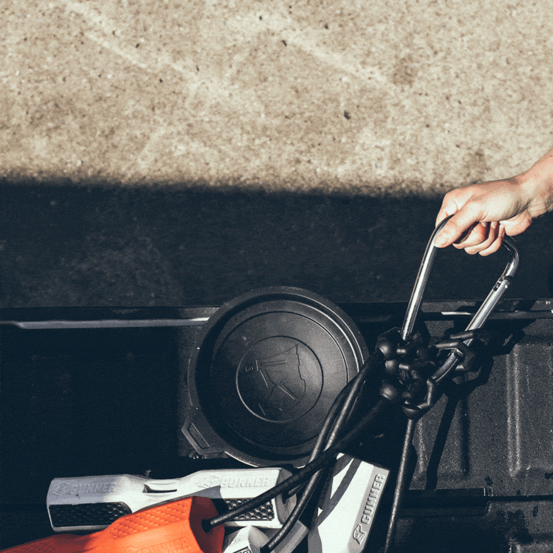A person’s hand holds the handlebar of an orange and white STIHL leaf blower, part of essential outdoor gear, which is placed in the back of a black truck bed on a sunny day.